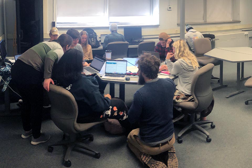 A group of statistics students and tutors work together at a round table. A male tutor in the foreground kneels on the floor while helping a female student on her laptop at the table.